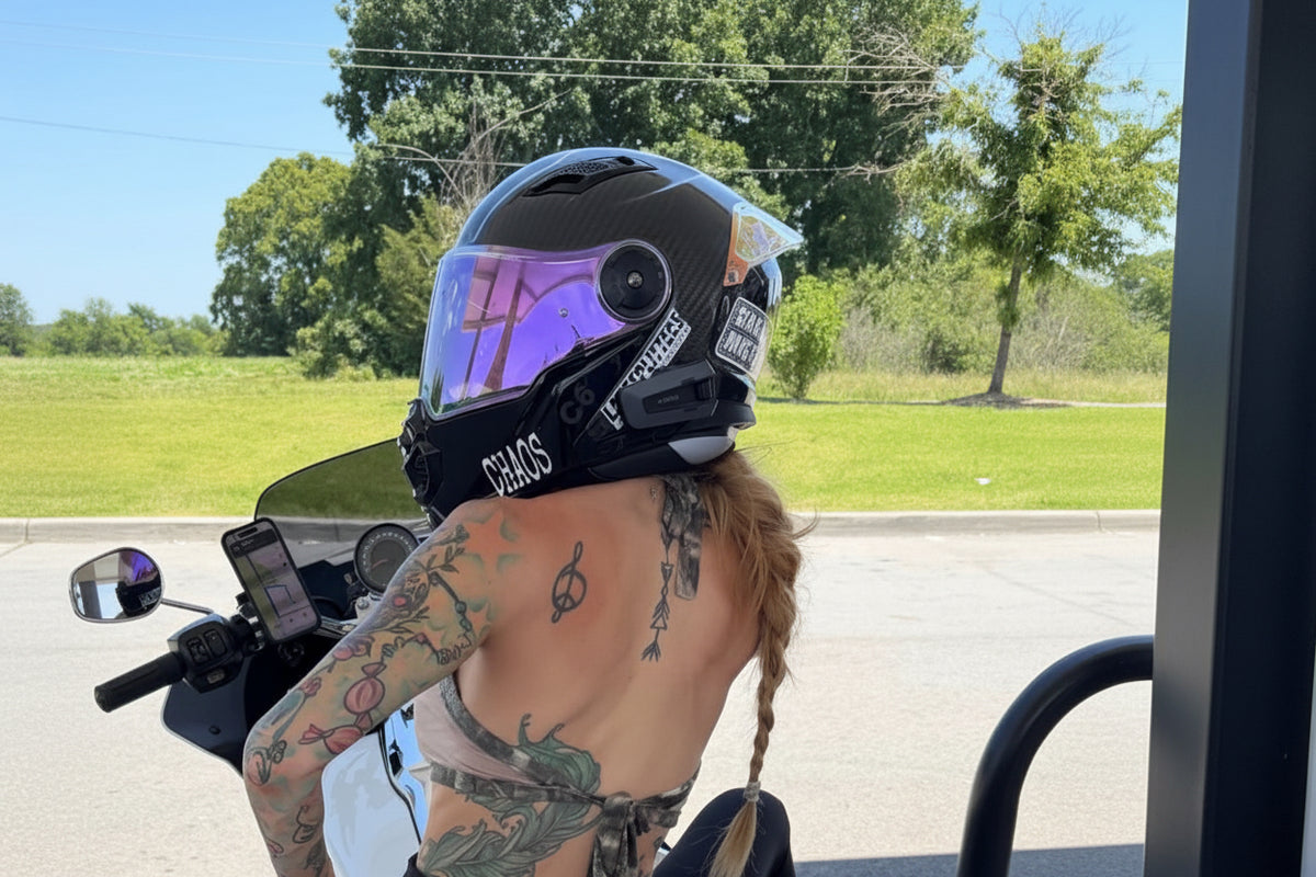 A woman with tattoos sits on a white and black three-wheeled motorcycle outdoors, wearing a black full-face helmet with a reflective visor.